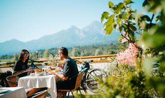 A couple is sitting outside at a table enjoying their breakfast. In the background, mountains and a beautiful landscape can be seen.