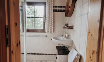 A cozy bathroom with a bright wooden ceiling and tiles. There is a shower, a sink, and a window with curtains.