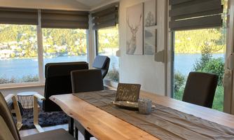 A cozy dining area with a large wooden table and black chairs. Through the windows, you can see a beautiful view of the lake and the surrounding landscape.
