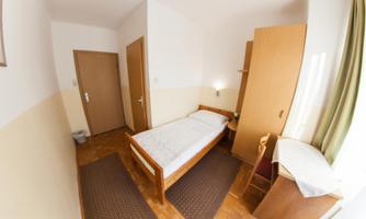 A simple room with a single bed, a desk, and a wardrobe. The walls are light, and there is a window with curtains.