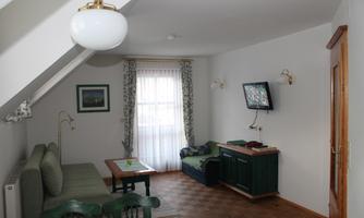 A cozy living and bedroom with a green sofa and a table. A television and windows provide brightness in the room.