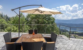 A cozy dining table on a terrace with a sunshade and a view of the mountains and the lake. The sky is blue with some clouds.