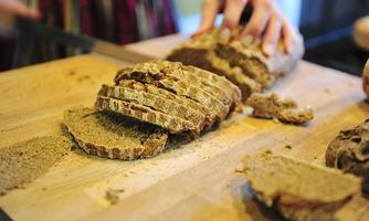 Freshly baked bread on a wooden board, being sliced. In the background, hands are visible holding the bread.