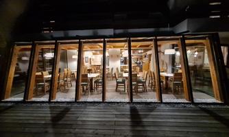 A cozy restaurant with large windows and wooden furniture. It is illuminated in the evening and offers an inviting atmosphere.