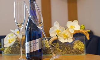 A bottle of Prosecco with two champagne glasses is on a table. In the background, elegant floral arrangements can be seen.