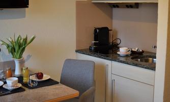 A small, modern kitchen with a table set for breakfast. A coffee machine and some drinks are on the countertop, while a plant is decoratively placed on the table.