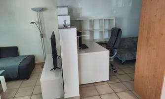 A simply furnished room with a desk, a television, and an office chair. On the left side, there is a sofa and a floor lamp.