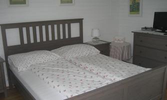 A cozy bedroom with a double bed and light-colored walls. There is a bedside lamp and a TV on a cabinet.
