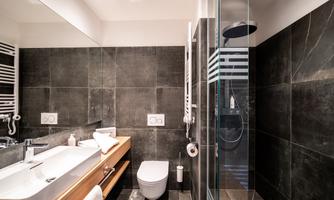 A modern bathroom with dark tiles and a glass shower. There is a wooden washstand and a wall-mounted toilet.