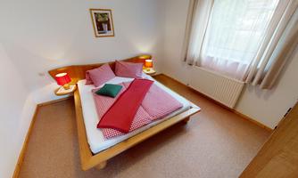 A cozy bedroom with a double bed, red pillows, and warm lamps. The windows let in plenty of daylight, giving the room an inviting atmosphere.