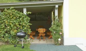 A cozy outdoor area with a wooden table and benches. In the foreground, there is a grill surrounded by plants.