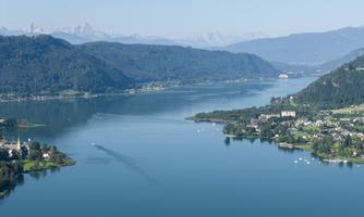 A picturesque landscape with a calm lake, surrounded by green hills and mountains in the background. The sky is clear and blue, highlighting the beauty of nature.
