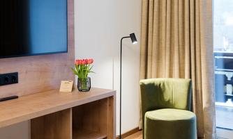 A modern room with a television, a table, and a small floor lamp. A green chair and a window with curtains complete the cozy atmosphere.