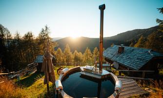 A cozy area with a wooden outdoor bath, surrounded by nature. In the background, the sun shines over the mountains.