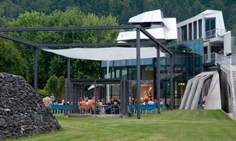 A modern building with large windows and a unique design. In the foreground, an outdoor event is taking place with guests at tables.