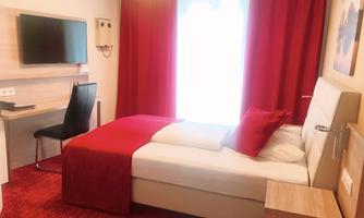 A modern hotel room with a single bed and red bed linen. The room features a desk, a chair, and a television.