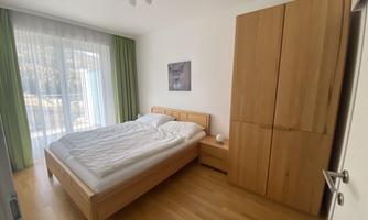 A bright bedroom with a double bed and wooden furniture. Large windows with curtains let in plenty of light.