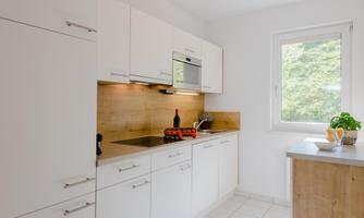 A modern kitchen with white cabinets and wooden accents. The space is bright and inviting, with a large window and a pleasant atmosphere.