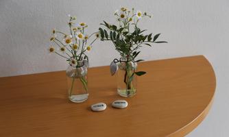 Two small vases with white flowers are on a wooden shelf. In front of the vases are three stones with the inscriptions "Believe," "Hope," and "Love."