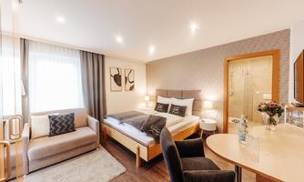 A modern hotel room with a double bed, a sofa, and a table. The decor is elegant and bright, with soft colors and ample light.