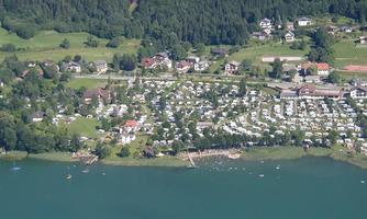 A picturesque landscape with a clear lake and many holiday homes. In the background, green meadows and wooded hills can be seen.