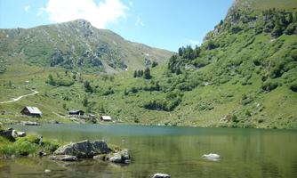 A tranquil mountain lake surrounded by green hills and forests. In the background, some huts are visible.