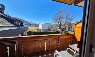 A balcony with a view of the mountains and blue sky. Next to it are a few trees and residential houses.