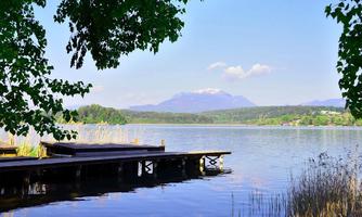 A tranquil lake landscape with green trees and a wooden walkway. In the background, mountains and a blue sky can be seen.
