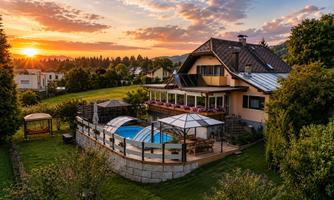 A beautiful house with a pool and a terrace, surrounded by green meadows. In the background, the sunset illuminates the sky.