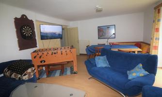 A cozy living room with blue sofas and a foosball table. A TV hangs on the wall and a dartboard is also available.