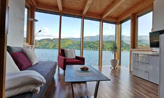 A modern living room with large windows and a view of the lake. The décor is bright and inviting, featuring a sofa, an armchair, and a table.