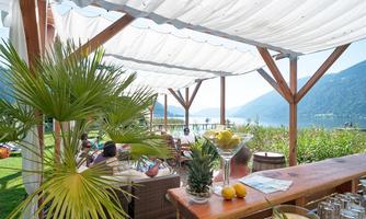 A cozy outdoor area with a view of a lake and surrounding mountains. Plants and a wooden bar table lend the scene a relaxed atmosphere.
