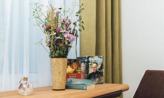 A vase with colorful dried flowers is on a wooden table. Next to it lie some books and a decorative glass vase.