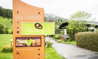 A welcome sign of the Mauerwirt shows the arrival at an inn in a picturesque landscape. Surrounded by green hills and trees, it is an inviting place for visitors.