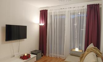 A modern living room with a television on the wall and a comfortable sofa. The windows are decorated with white curtains and red drapes.