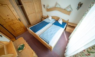 A cozy bedroom with a wooden bed and blue bedding. The room has a wardrobe and a desk with a window that lets in daylight.