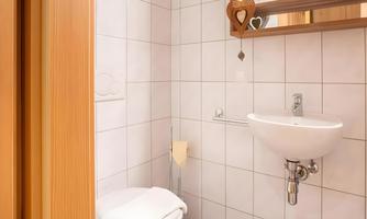 A small, modern bathroom with white tiles. It includes a toilet, a small sink, and a mirror.