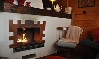 A cozy fireplace with a glowing fire in a wood room. Next to the fireplace is a wooden chair with a blanket.