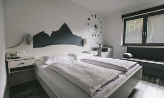 A modern bedroom with a large bed and a minimalist design. The walls are in neutral colors, and there is a stylish clock as well as a window with a view outside.