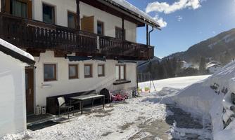 A cozy house in the snow with a balcony and outdoor seating. In the background, there are snow-covered mountains under a clear blue sky.