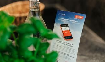 A glass bottle is next to a plant. An information sheet about the app "My Nassfeld Buddy" is on a table.