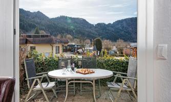 A cozy balcony with a table and chairs, surrounded by green plants. In the background, mountains and a clear sky are visible.