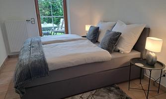 A cozy bedroom with a large bed, soft pillows, and a nightstand. Through the window, you can see a small garden area.