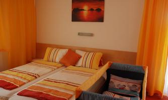 A cozy bedroom with two single beds and a crib. The walls are decorated with orange curtains and a picture of a sunset hangs above.
