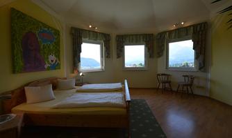 A bright room with two beds and large windows. A colorful artwork hangs on the wall, and there are wooden chairs and plants visible in the room.