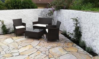 A cozy terrace with a seating area made of rattan furniture and a stone table. Surrounded by a white wall and some greenery.