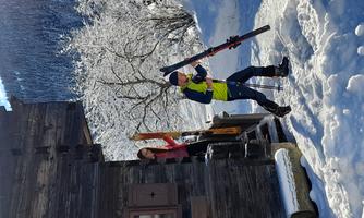 A skier is carrying his skis on the way to the snow. In the background, there is a wooden house surrounded by snow-covered trees.