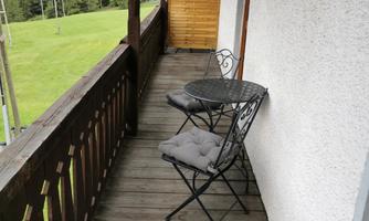 A cozy balcony with two chairs and a small table. In the background, green meadows and mountains can be seen.