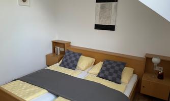A cozy bedroom with a double bed and two pillows. The walls are painted light, and there is a small bedside lamp.