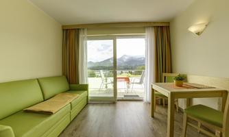 A modern room with a green sofa and a dining table. The windows offer a beautiful view of the mountains.